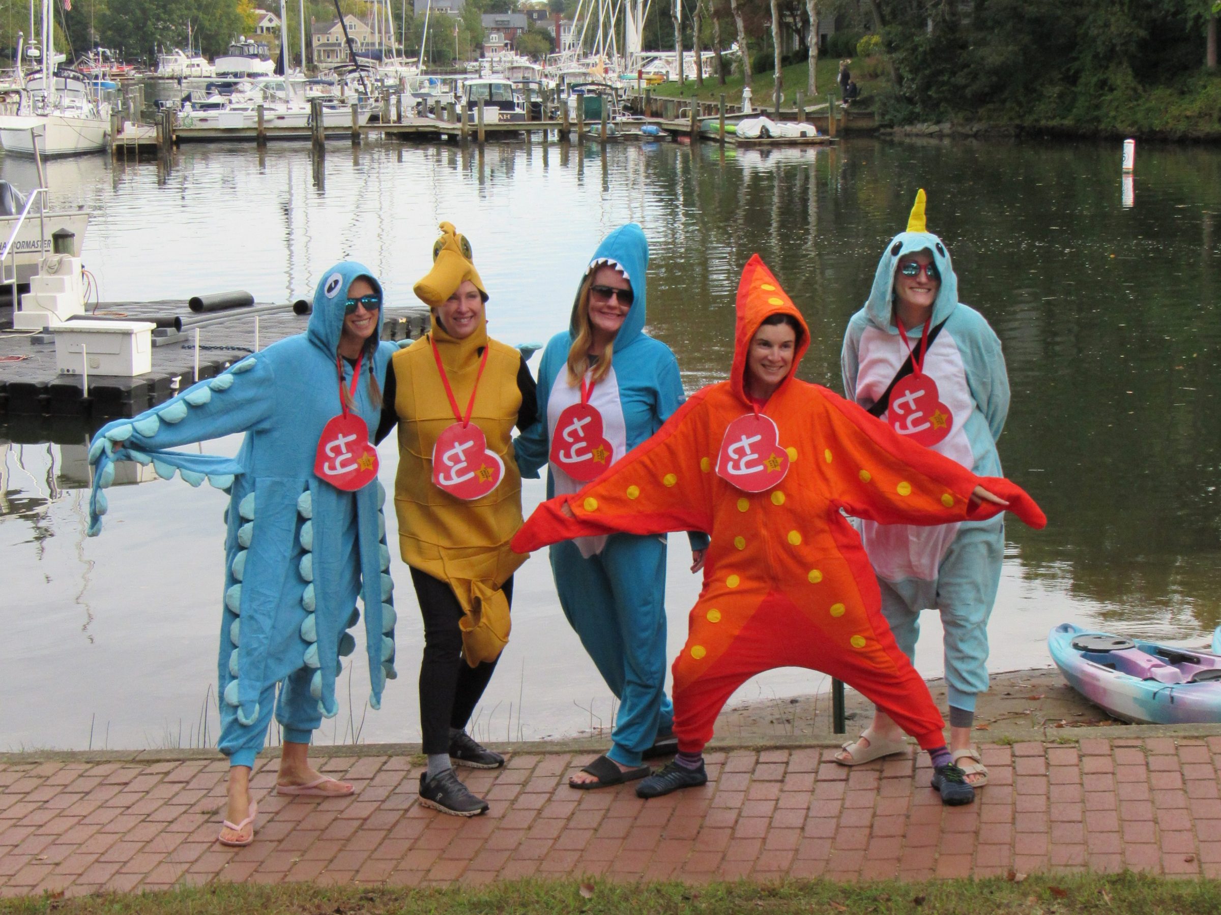 Five people in colorful sea creature costumes by a marina with boats and trees in the background.