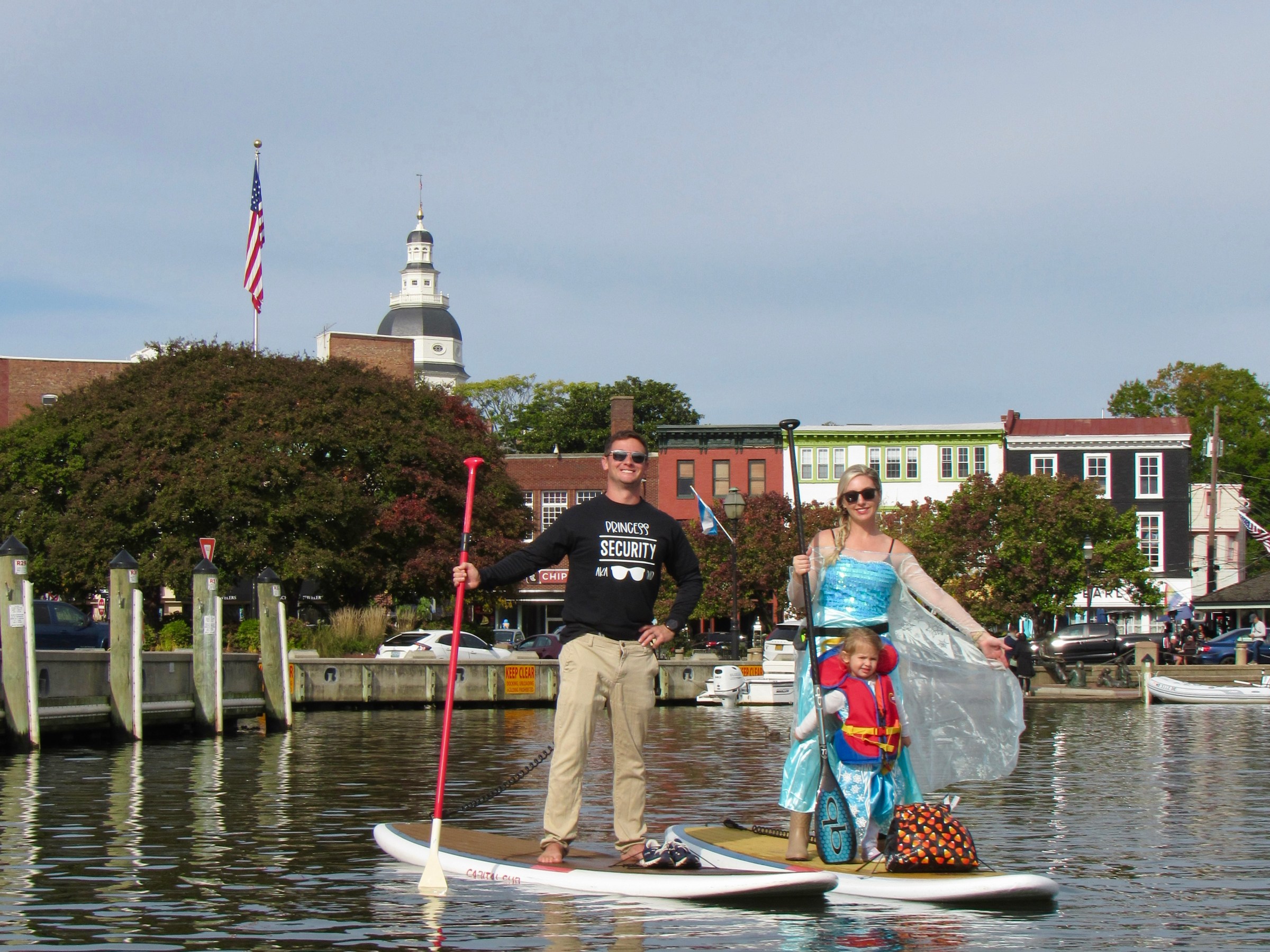 Two people paddleboarding, one in a security shirt and the other in a blue costume, on a calm waterfront.