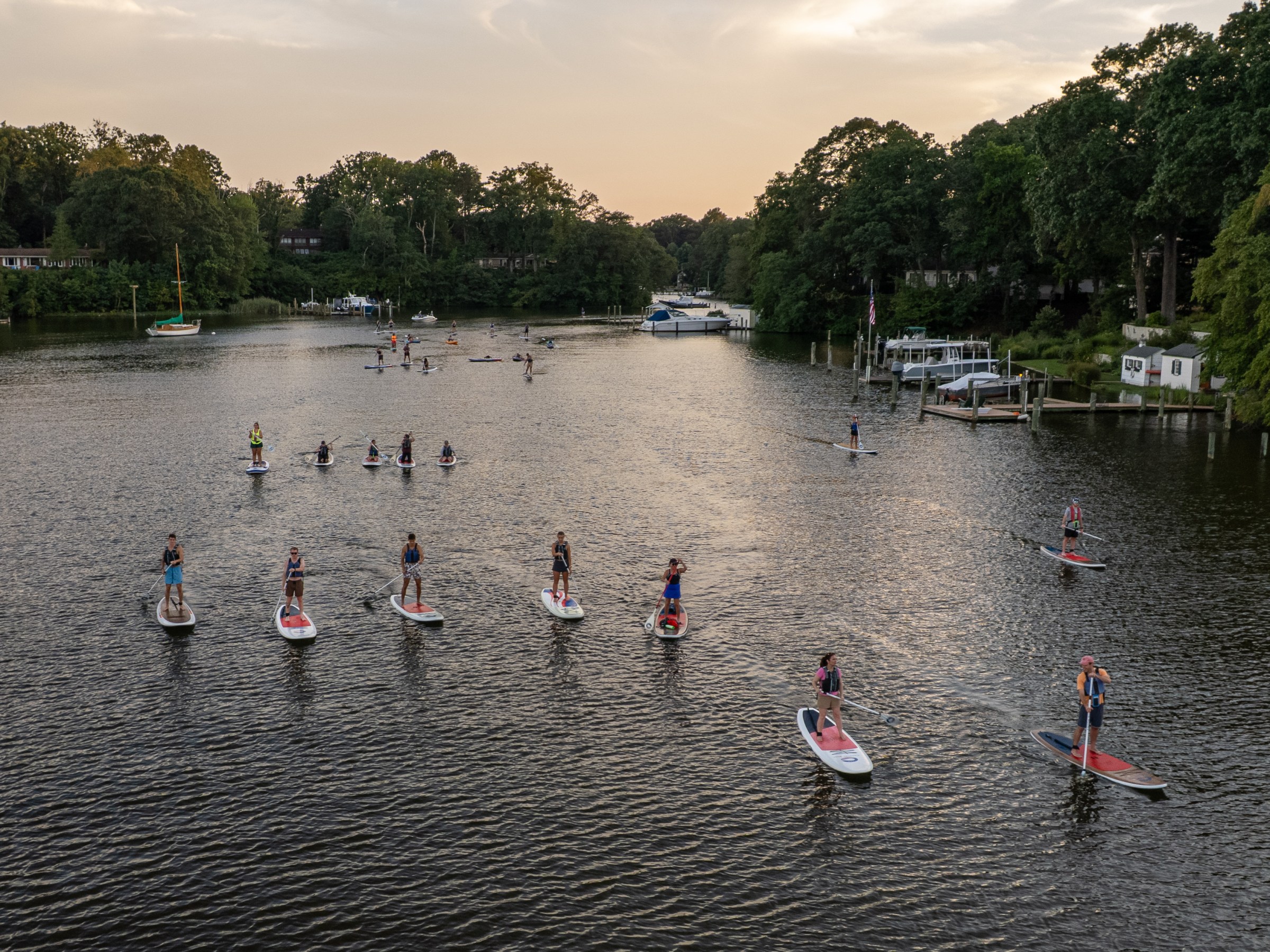 Group paddleboarding on a calm river with trees on both sides at sunset.