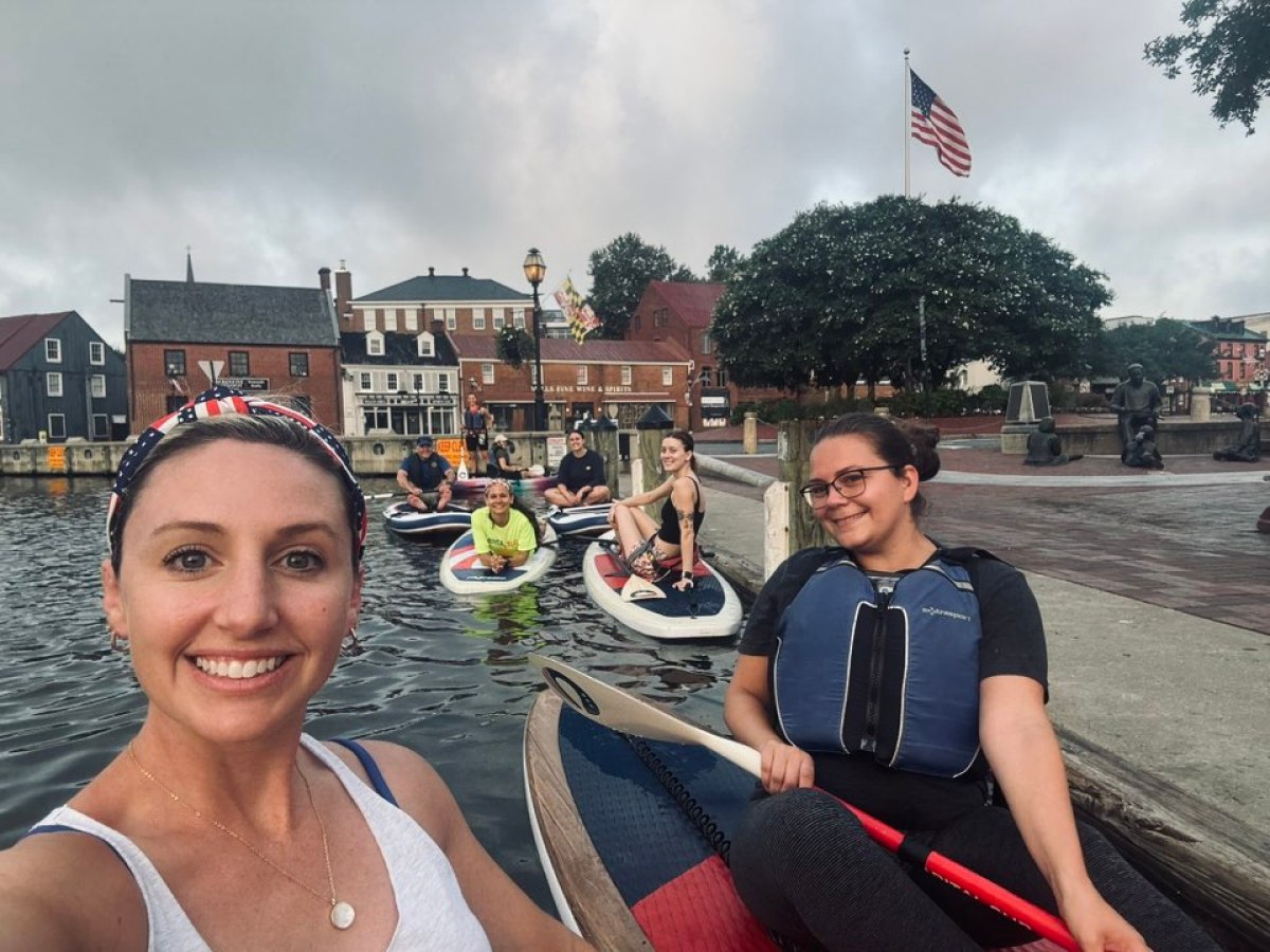 Group of people kayaking on a river with buildings and an American flag in the background.