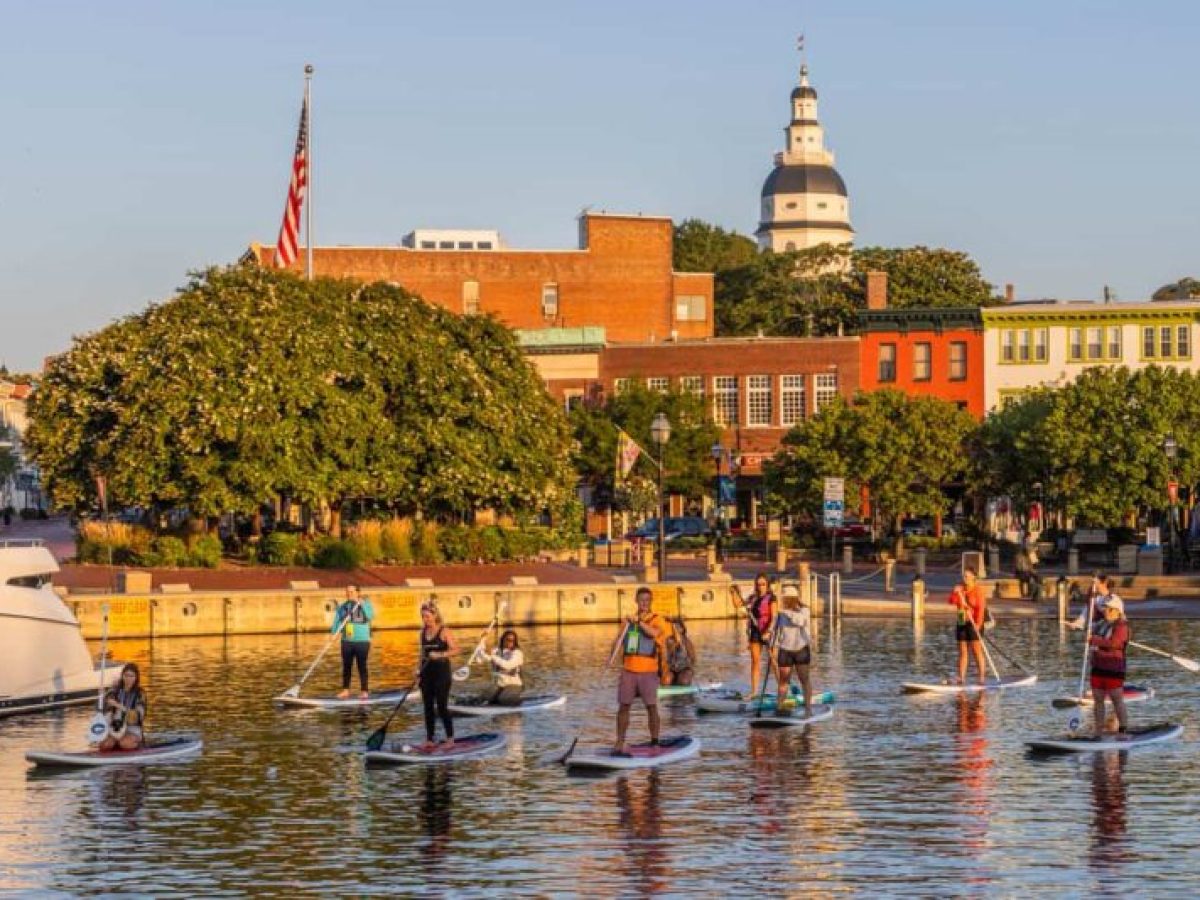 Group of people paddleboarding on water in front of colorful buildings and trees.