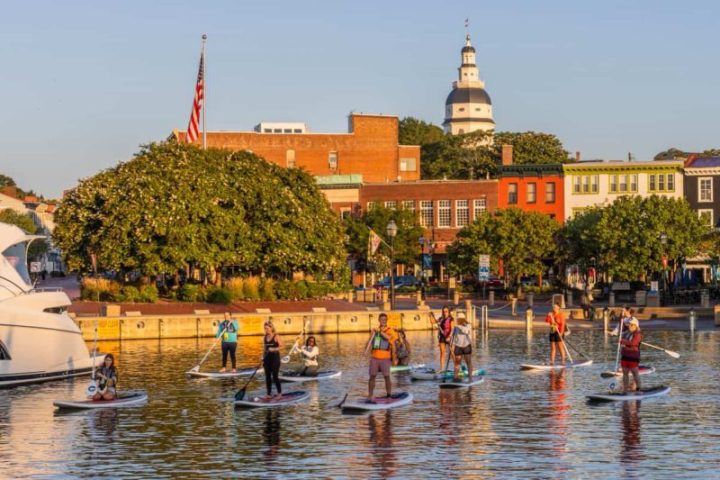 Group of people paddleboarding on water in front of colorful buildings and trees.