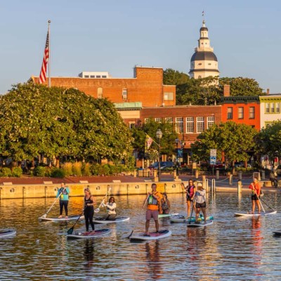 Group of people paddleboarding on water in front of colorful buildings and trees.