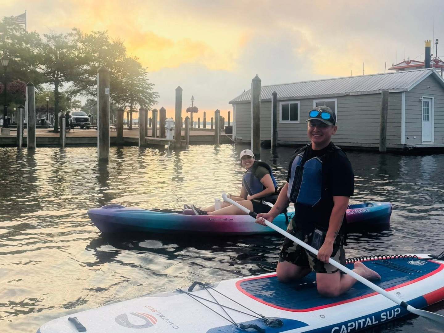 Two people on a paddleboard and kayak near a dock at sunset.