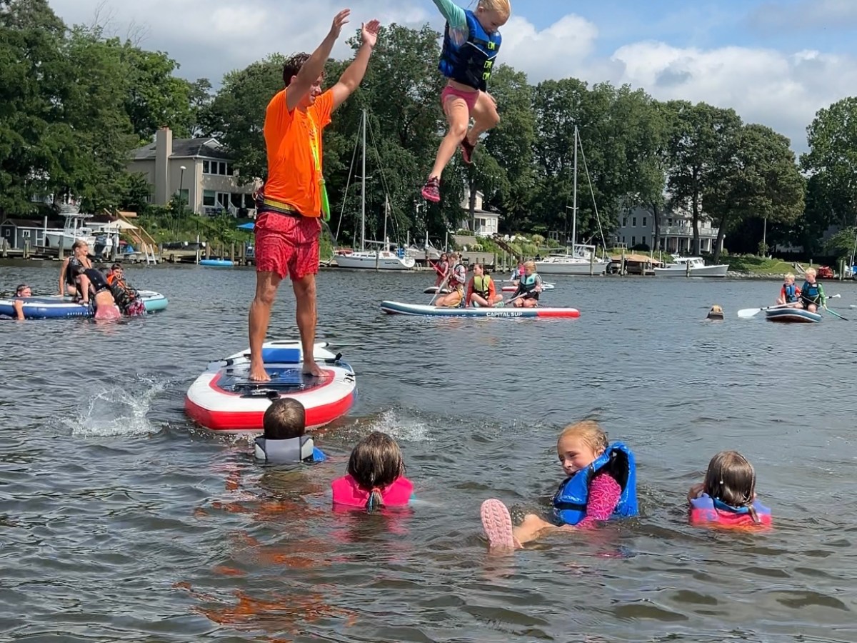 Man helps child jump off paddleboard into water; kids in life vests watching nearby.