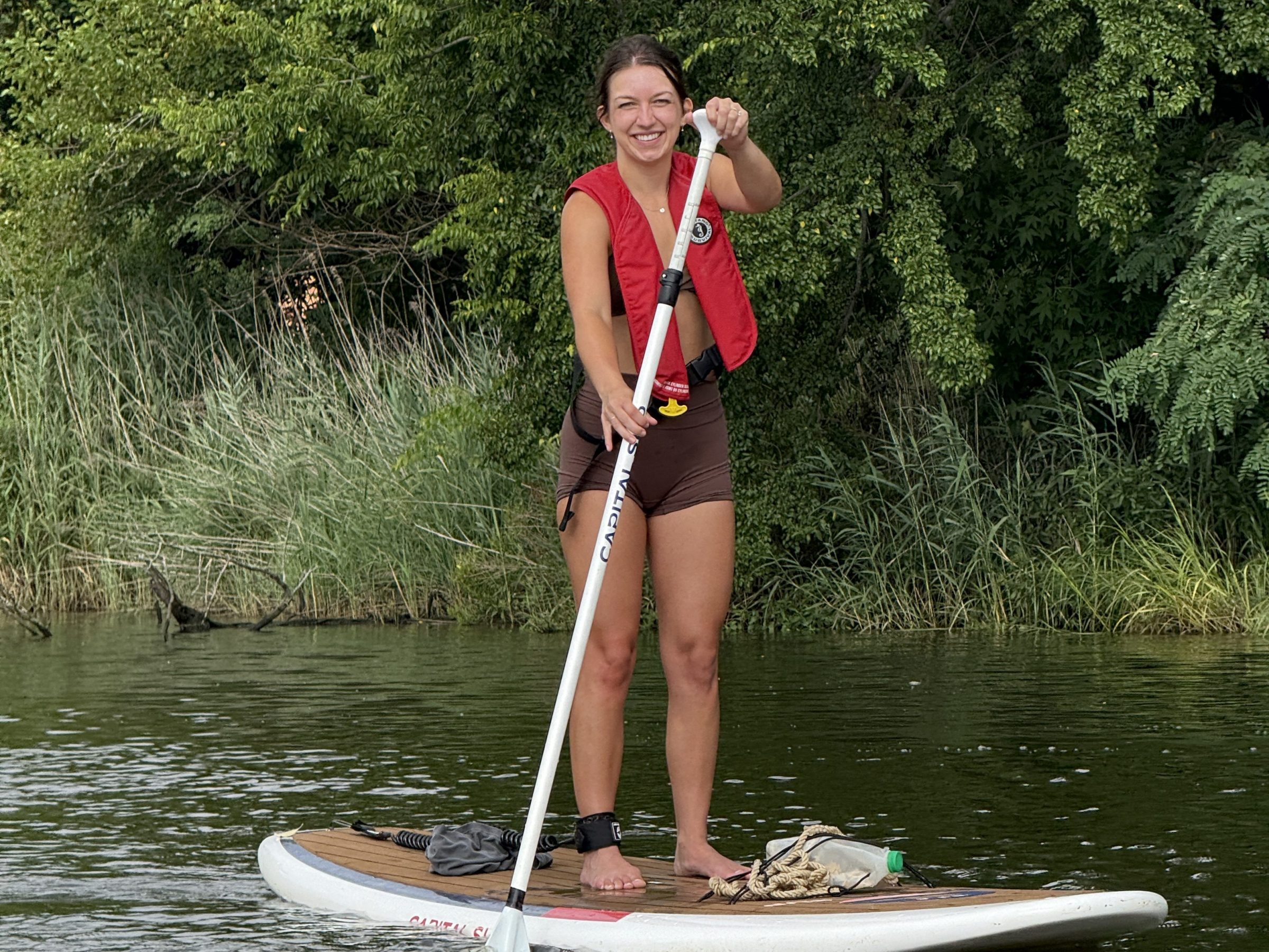 Person paddleboarding on a lake, wearing a red life jacket, with trees in the background.