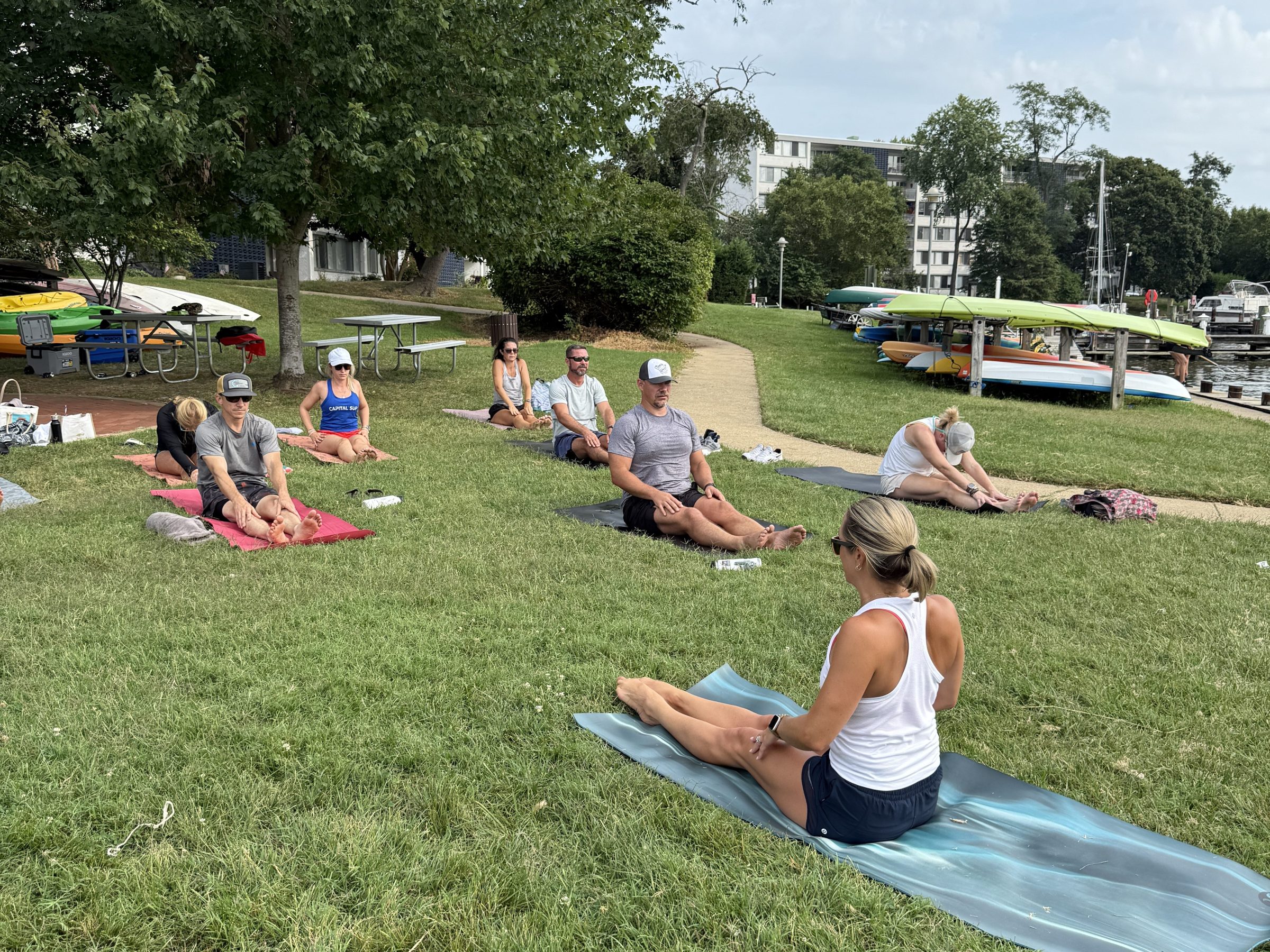 Outdoor yoga class on grass near water with trees and boats in background.