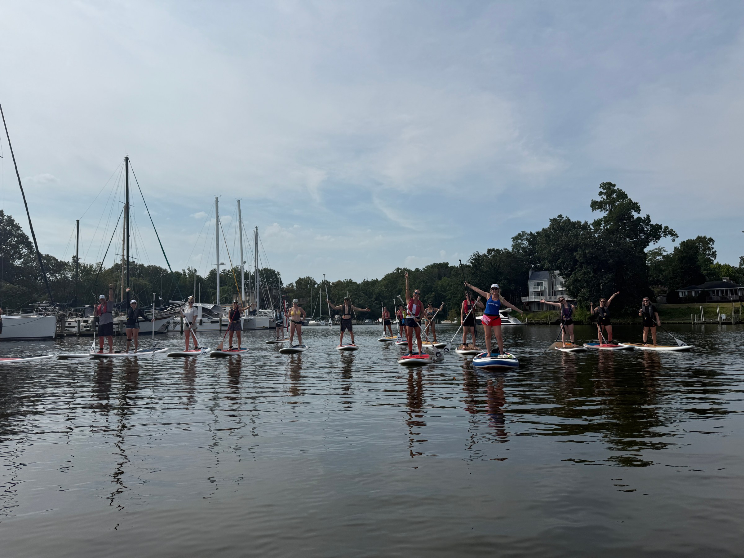 Group paddleboarding on calm water near sailboats under a cloudy sky.
