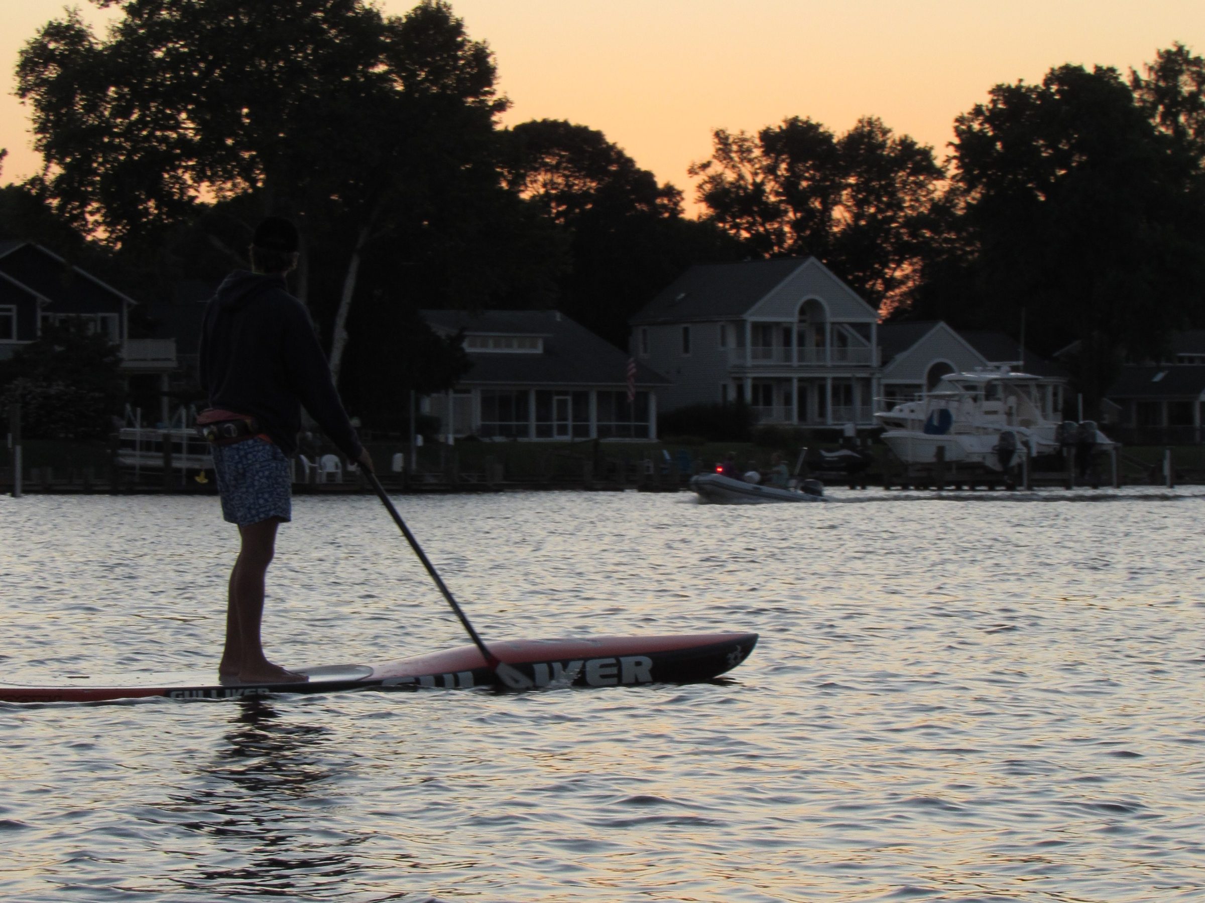 Person paddleboarding on a calm river at sunset, with houses and trees in the background.