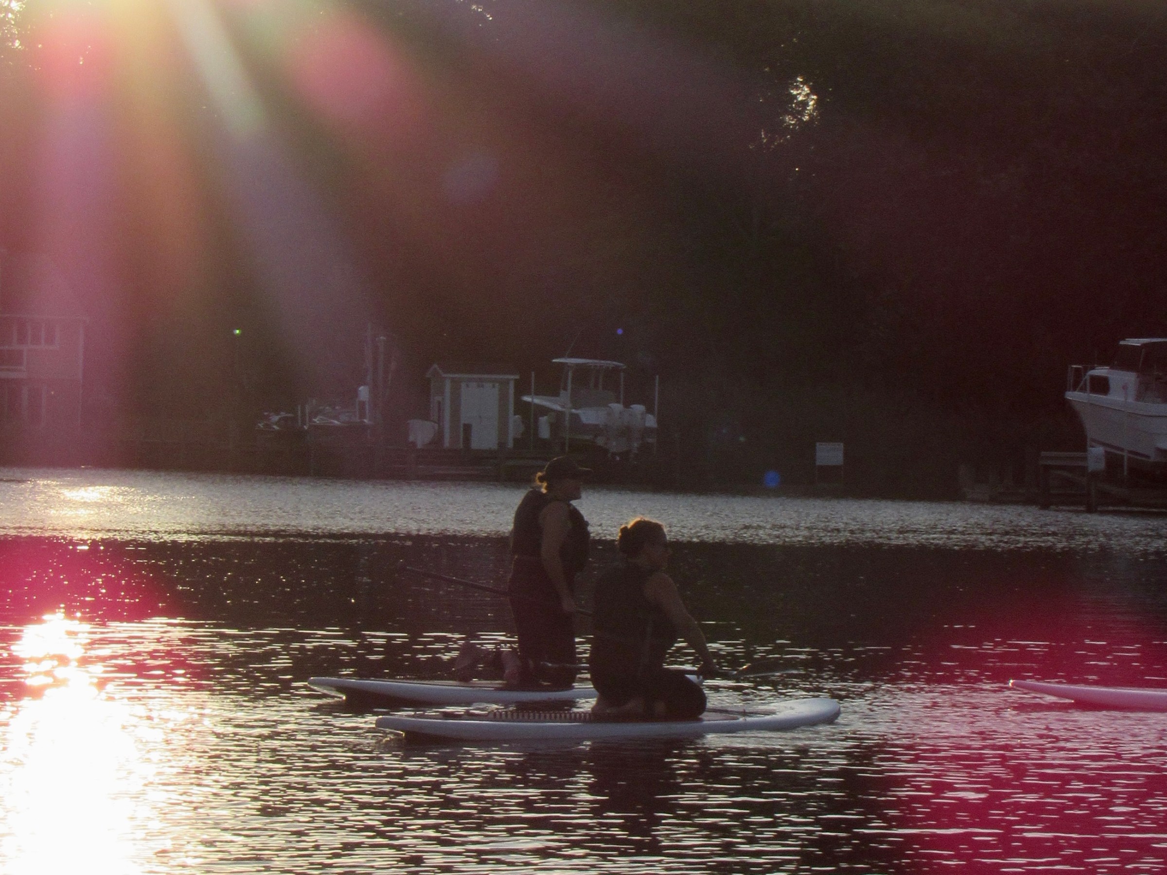 Two people paddleboarding on a calm lake at sunset with trees in the background.