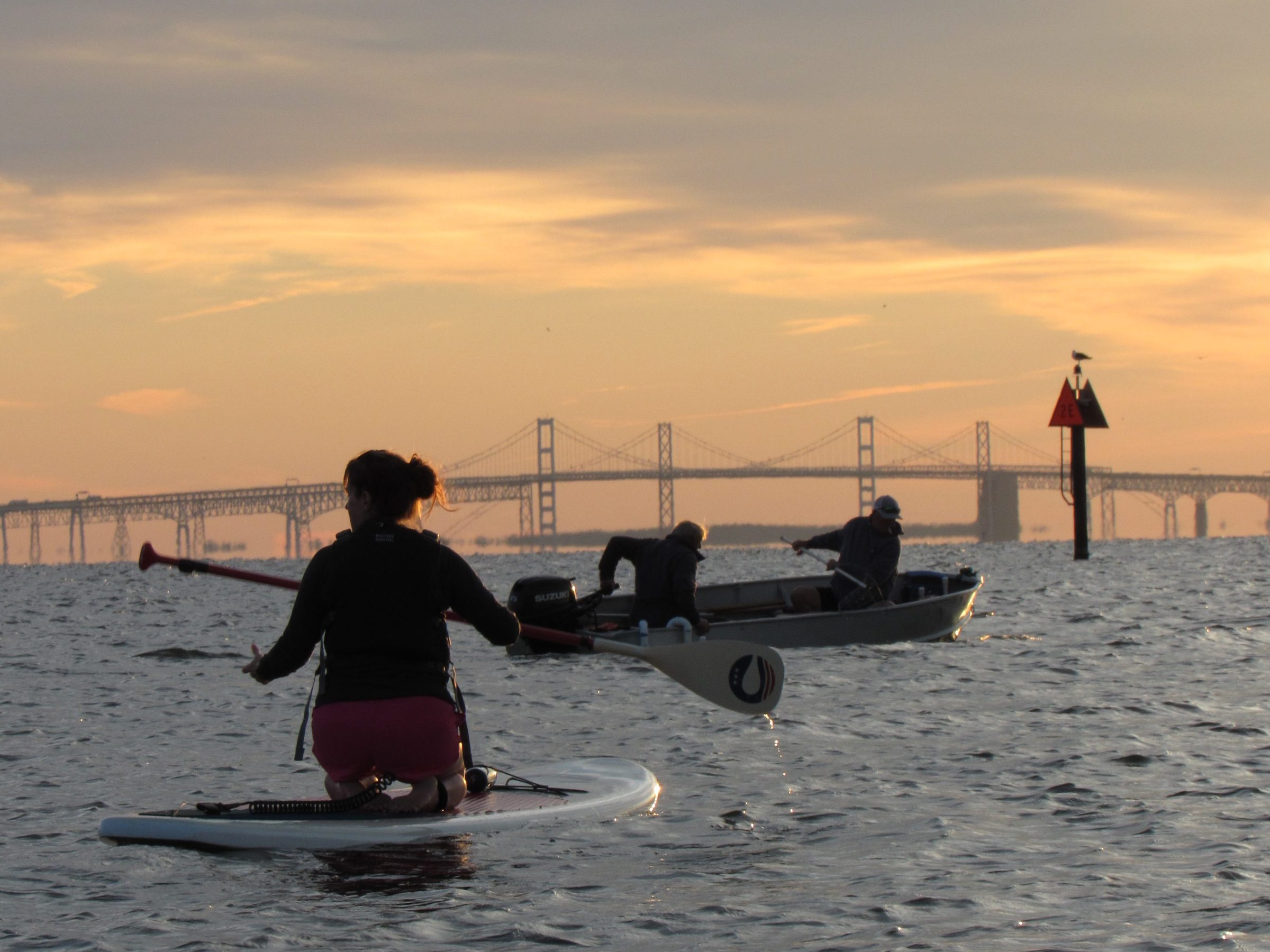 Person paddleboarding and two people in a boat on a calm river with a bridge in the background at sunset.