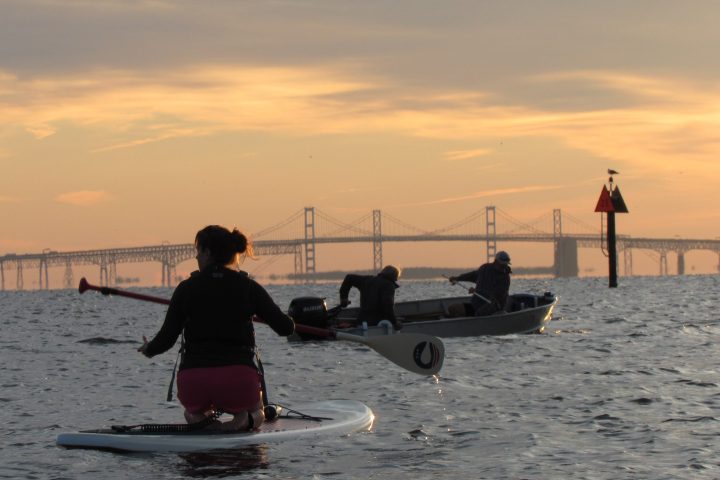 Person paddleboarding and two people in a boat on a calm river with a bridge in the background at sunset.