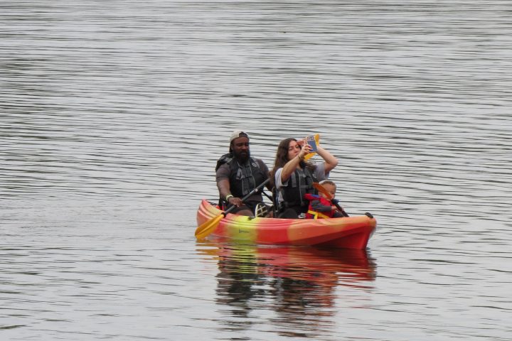 Three people in life jackets kayaking on a calm lake taking a selfie.