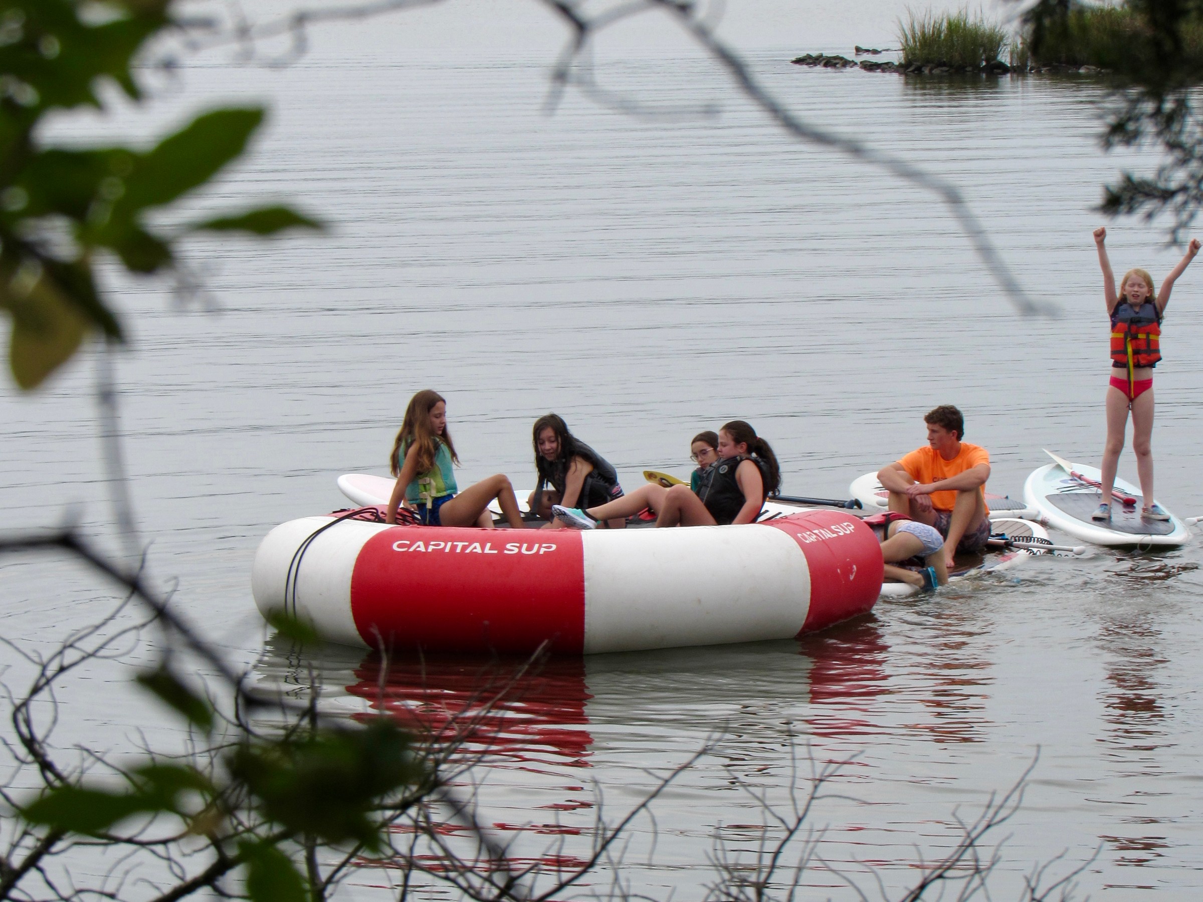 People on an inflatable platform in a lake, with trees in the foreground and houses in the background.