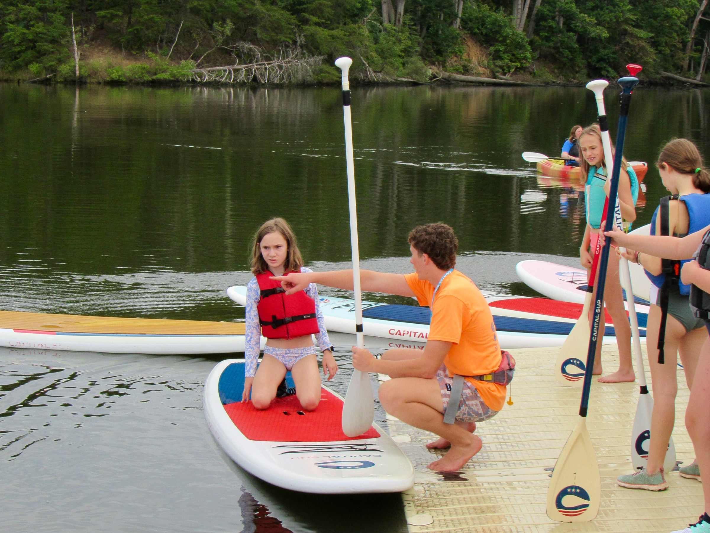 People on a dock preparing to paddleboard on a calm lake with trees in the background.
