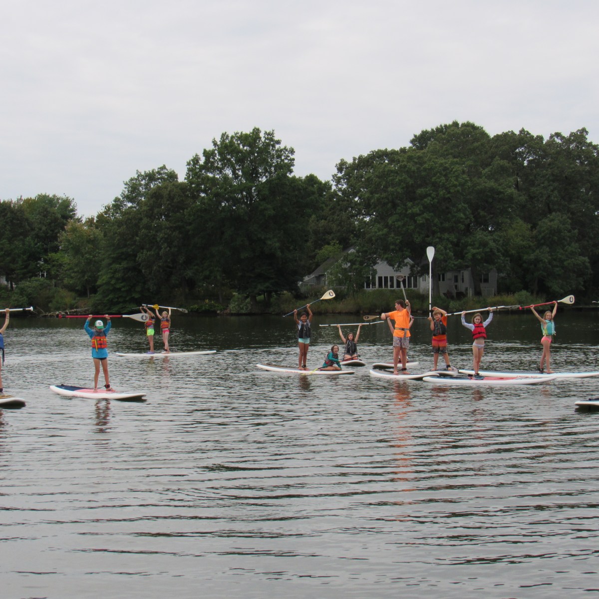 Group of people paddleboarding on a lake, holding paddles overhead, with trees in the background.