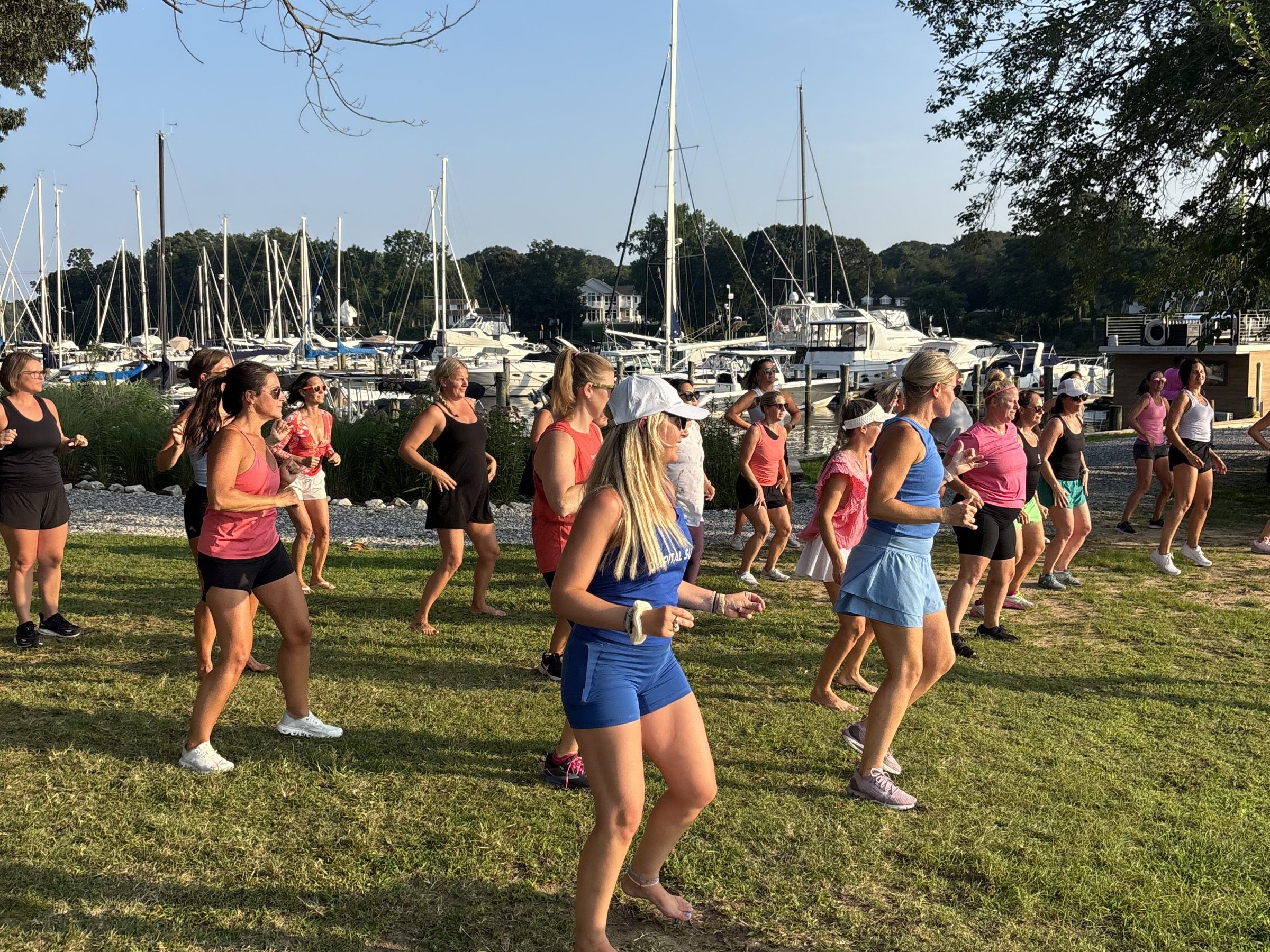 Group of people exercising outdoors near boats and trees on a sunny day.