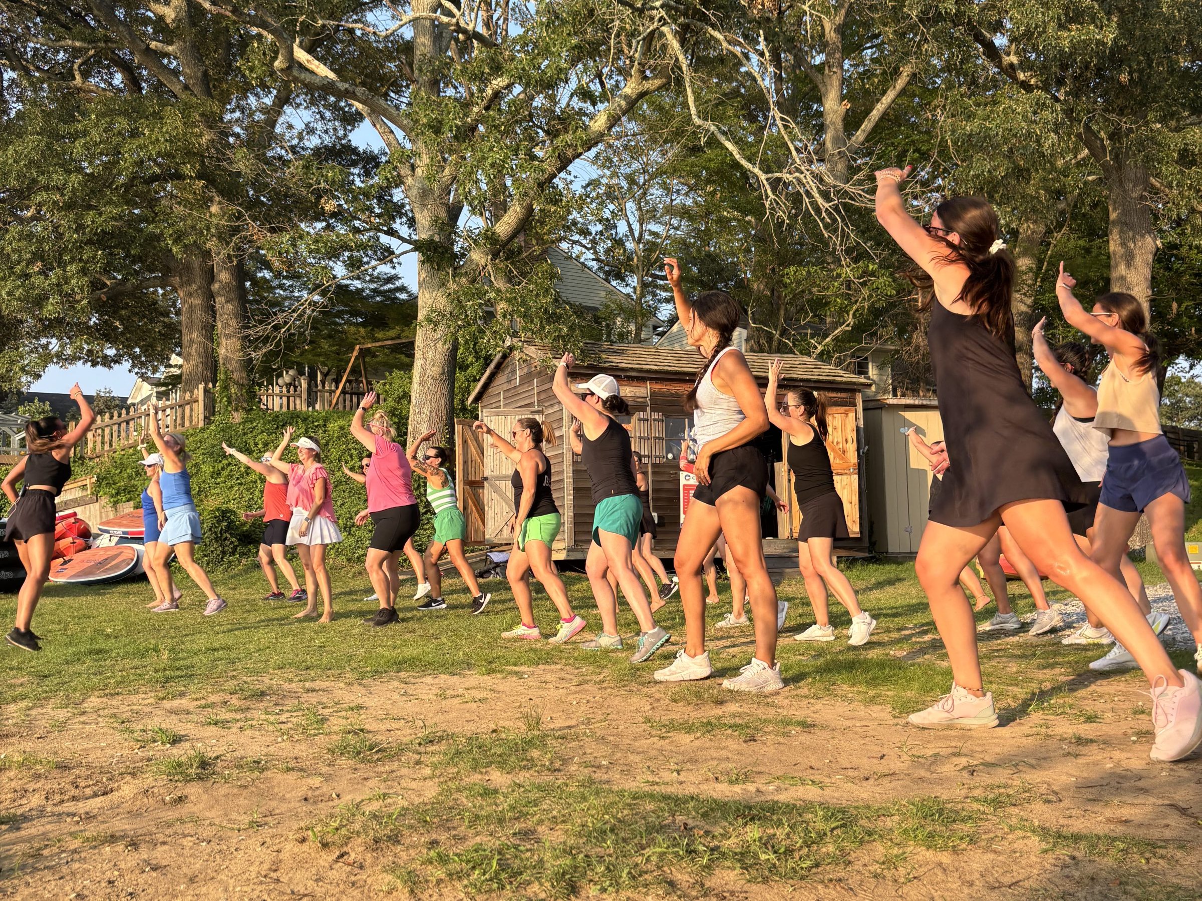 Group exercising outdoors, raising arms and moving on a grassy area with trees and sunlight.