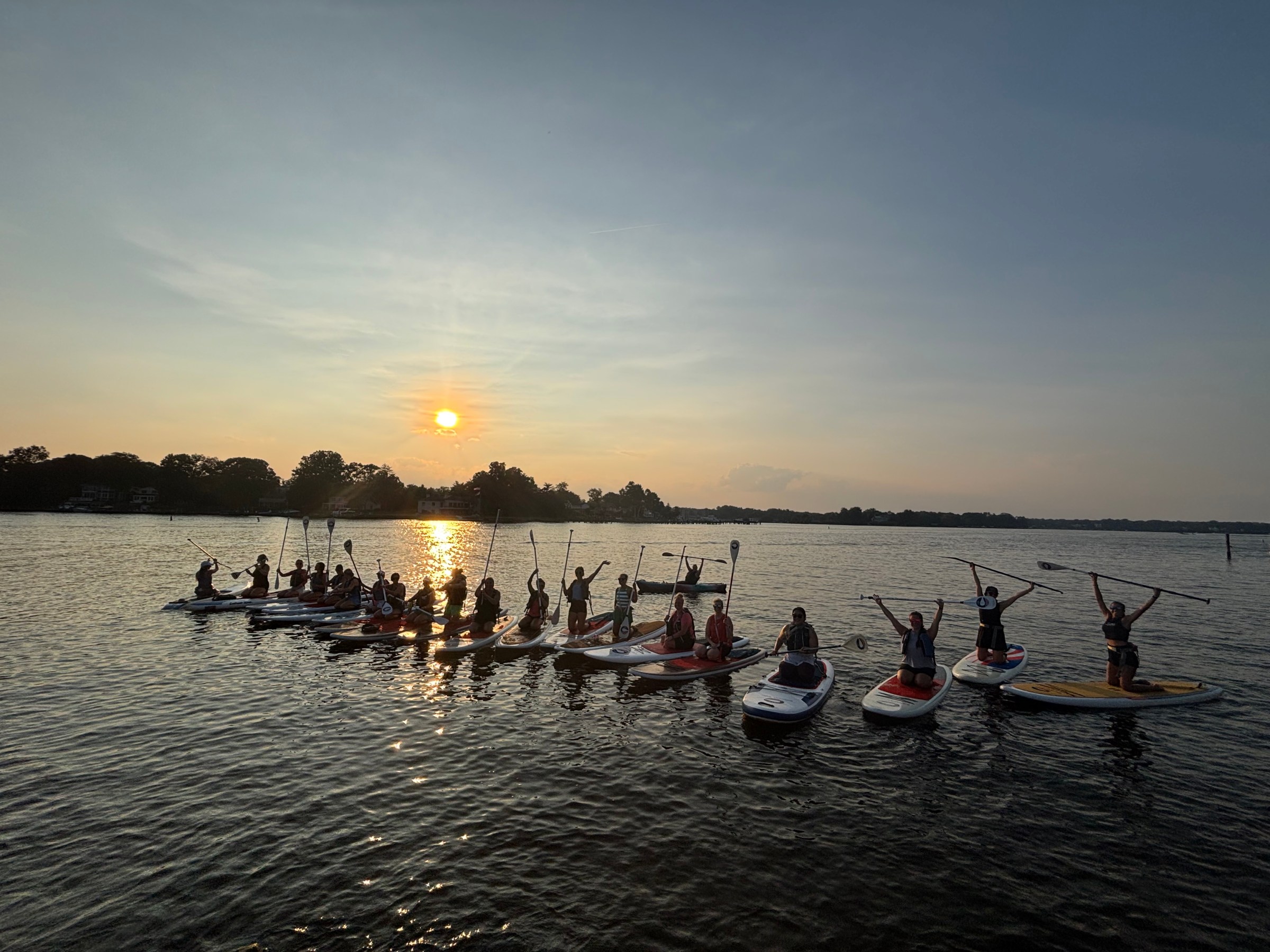 Group paddleboarding at sunset on a calm lake, paddles raised in celebration.