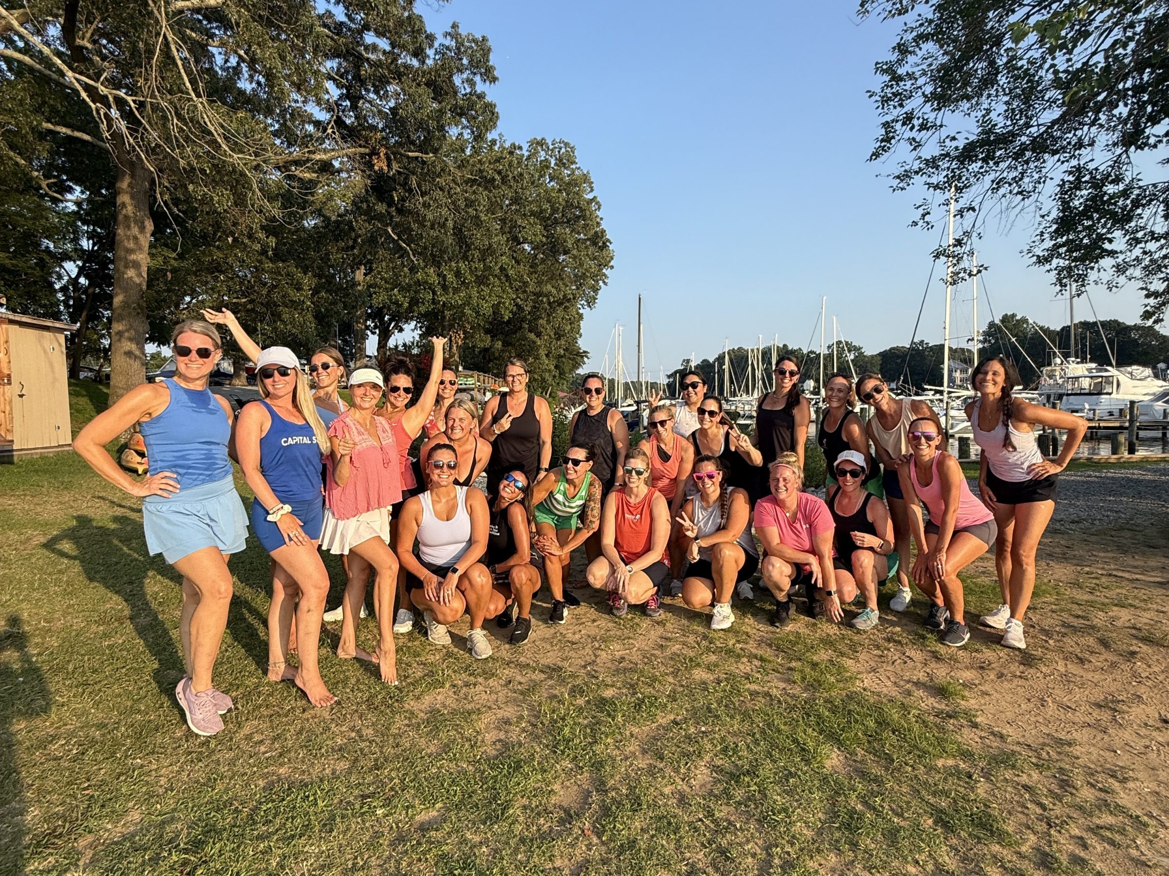 Group of people posing outdoors on grass with trees and boats in the background.