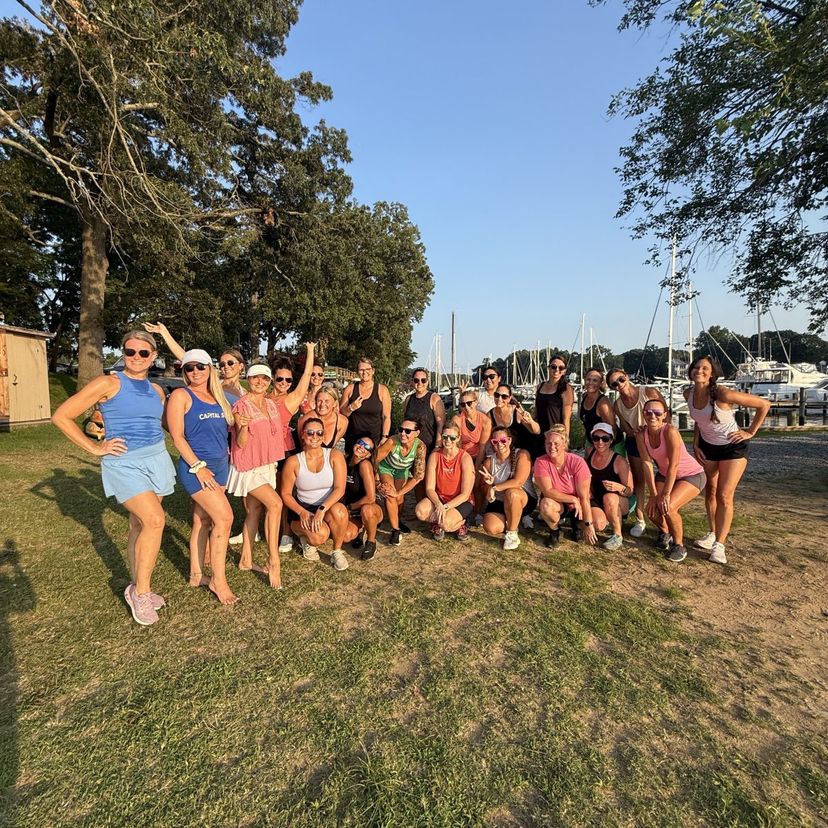 Group of people posing outdoors on grass with trees and boats in the background.