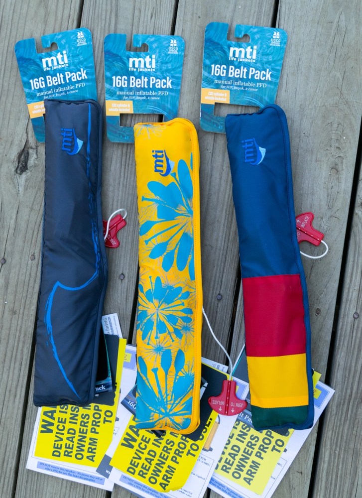 Three inflatable PFD belt packs in blue, yellow, and multicolor with packaging on wooden surface.