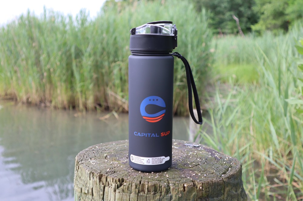 A black water bottle with logo on a wooden post by a pond, surrounded by tall grasses.