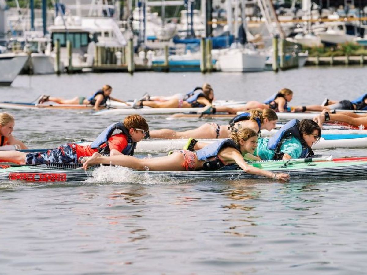 Children in life vests paddling on boards in a marina.