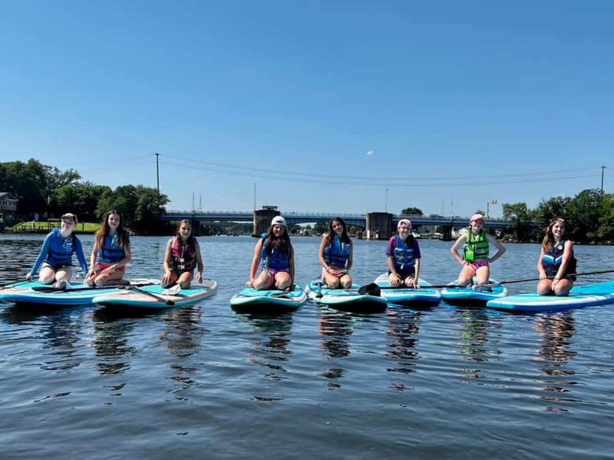 Seven people seated on paddleboards in a calm river under a clear blue sky.
