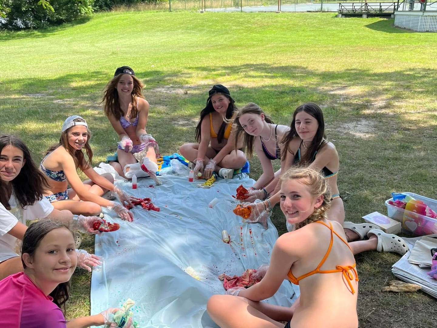 Group of girls outdoors making tie-dye shirts, sitting on grass with supplies.
