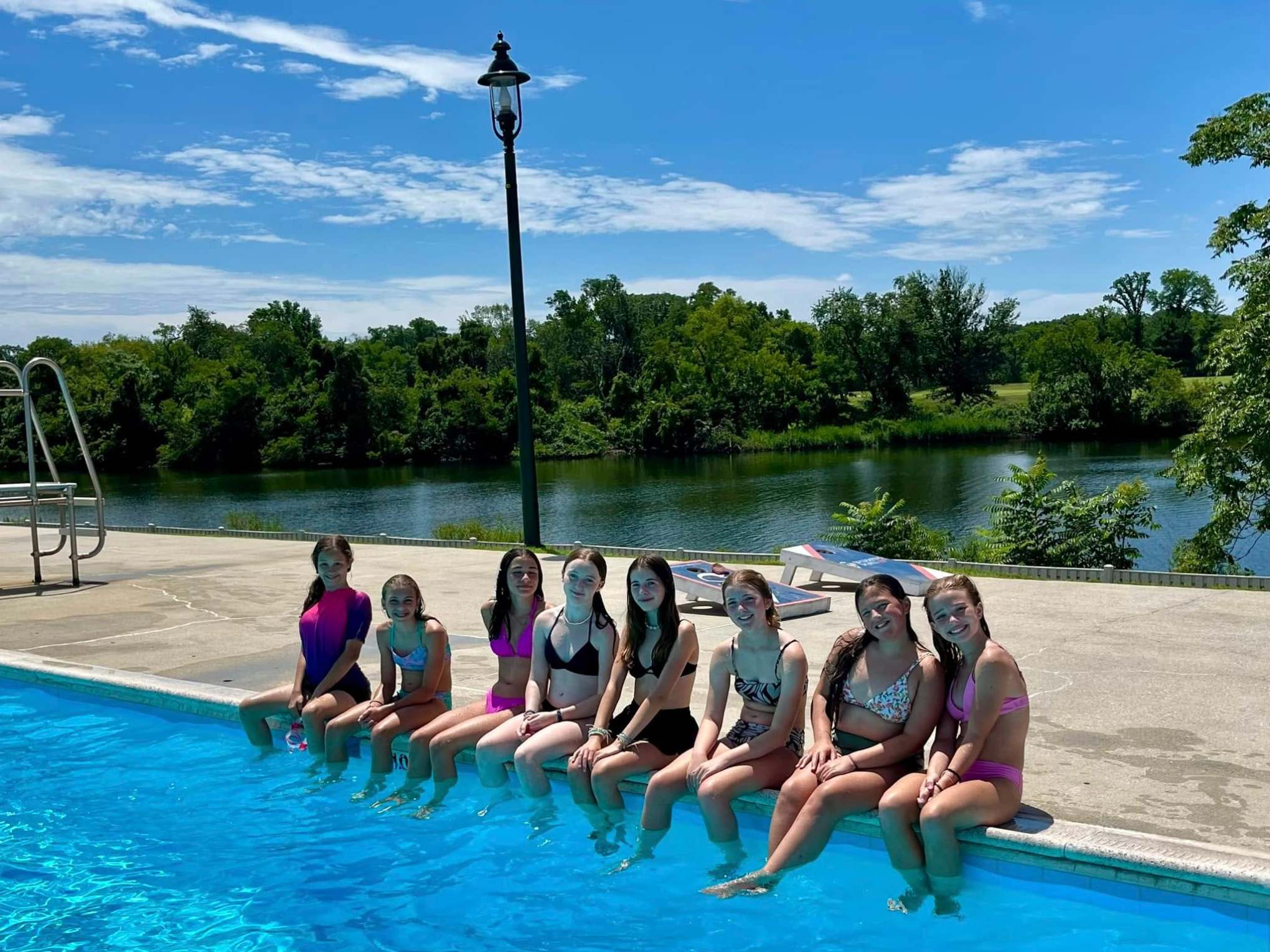 Eight girls sitting on the edge of a pool with a lake and trees in the background.