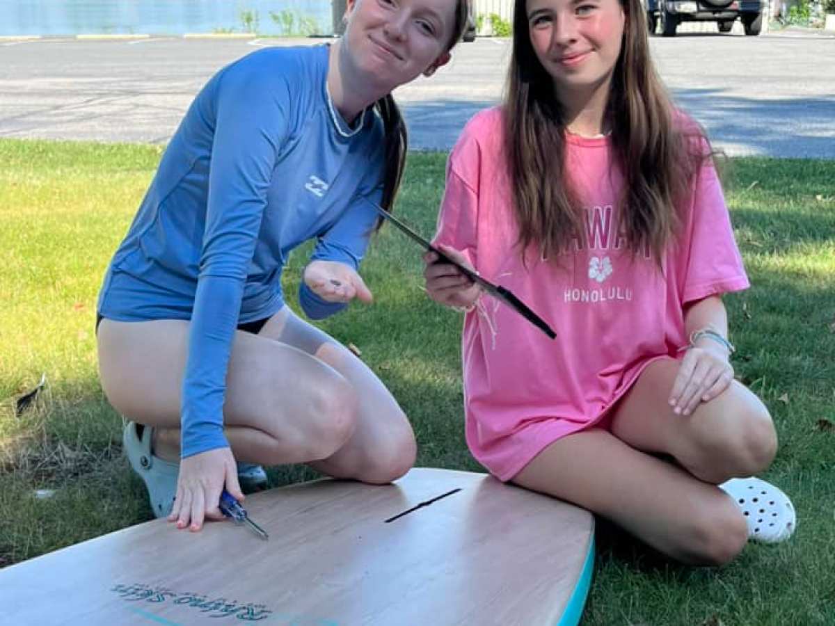 Two people kneeling on grass, holding tools near a surfboard, with water and a building in the background.