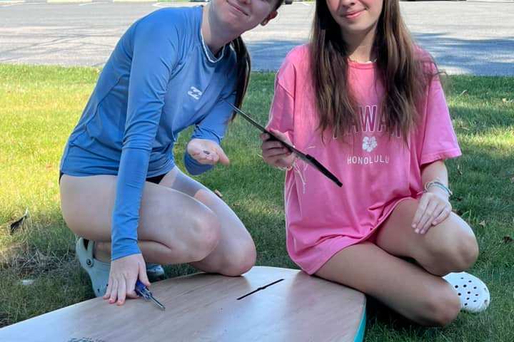Two people kneeling on grass, holding tools near a surfboard, with water and a building in the background.