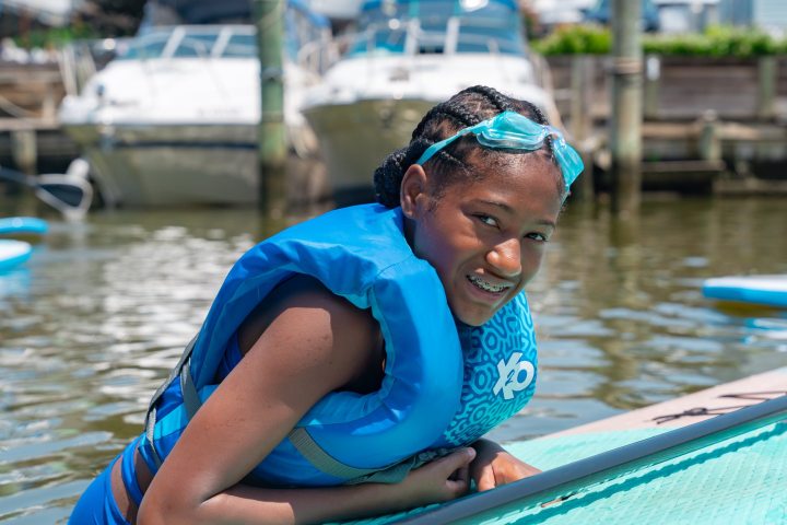 Child in life jacket and goggles smiling on paddleboard by boats.