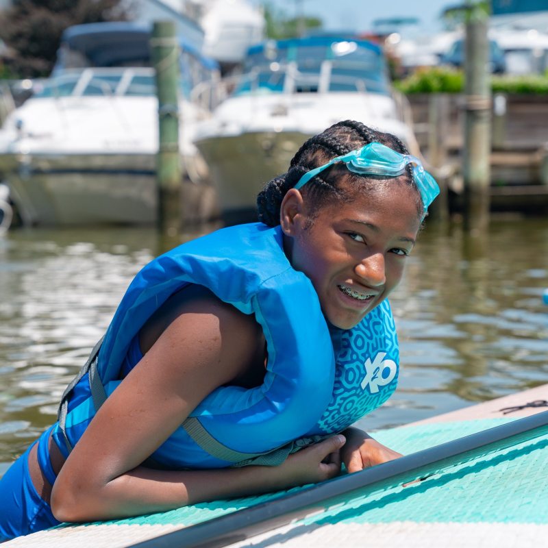 Child in life jacket and goggles smiling on paddleboard by boats.