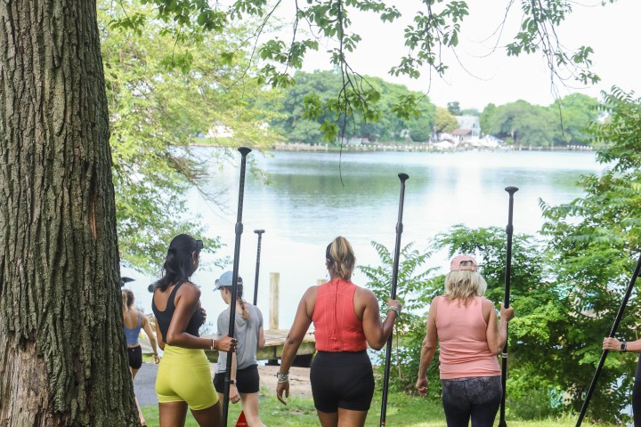 Group of people carrying paddles walking towards a lake under a tree.