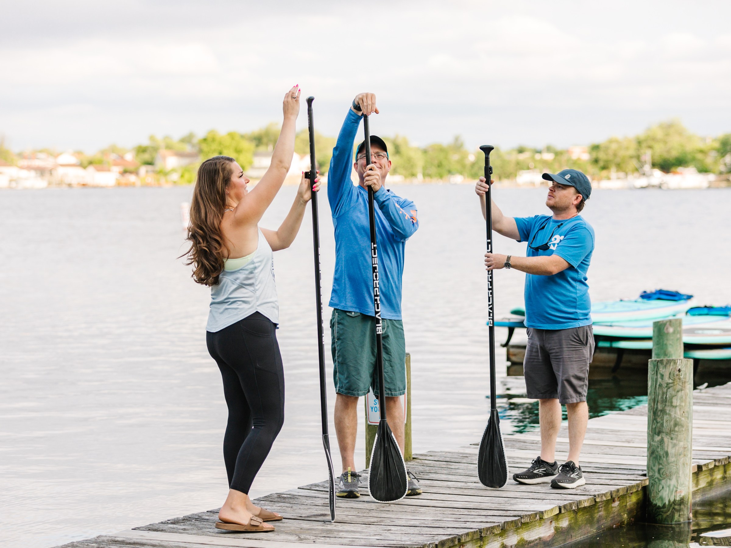 Three people adjusting stand-up paddleboard paddles on a lakeside dock.