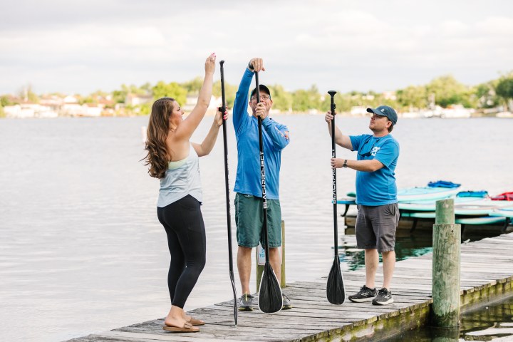 Three people adjusting stand-up paddleboard paddles on a lakeside dock.