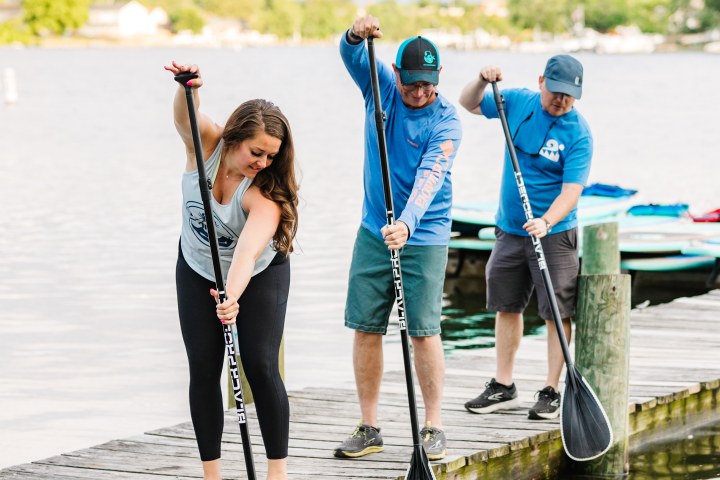 Three people on a dock practicing paddle strokes over calm water.