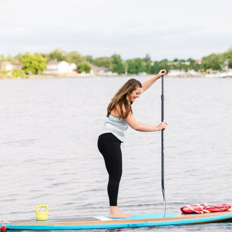 Person paddleboarding on a lake with a paddle and a kettlebell on the board.