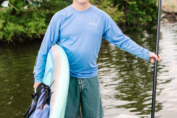 Man in blue shirt and cap holding paddleboard and paddle on dock by water.