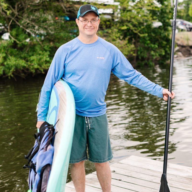 Man in blue shirt and cap holding paddleboard and paddle on dock by water.