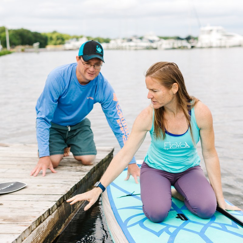 Two people at a dock, one kneeling on a paddleboard, the other on the dock, near calm water.