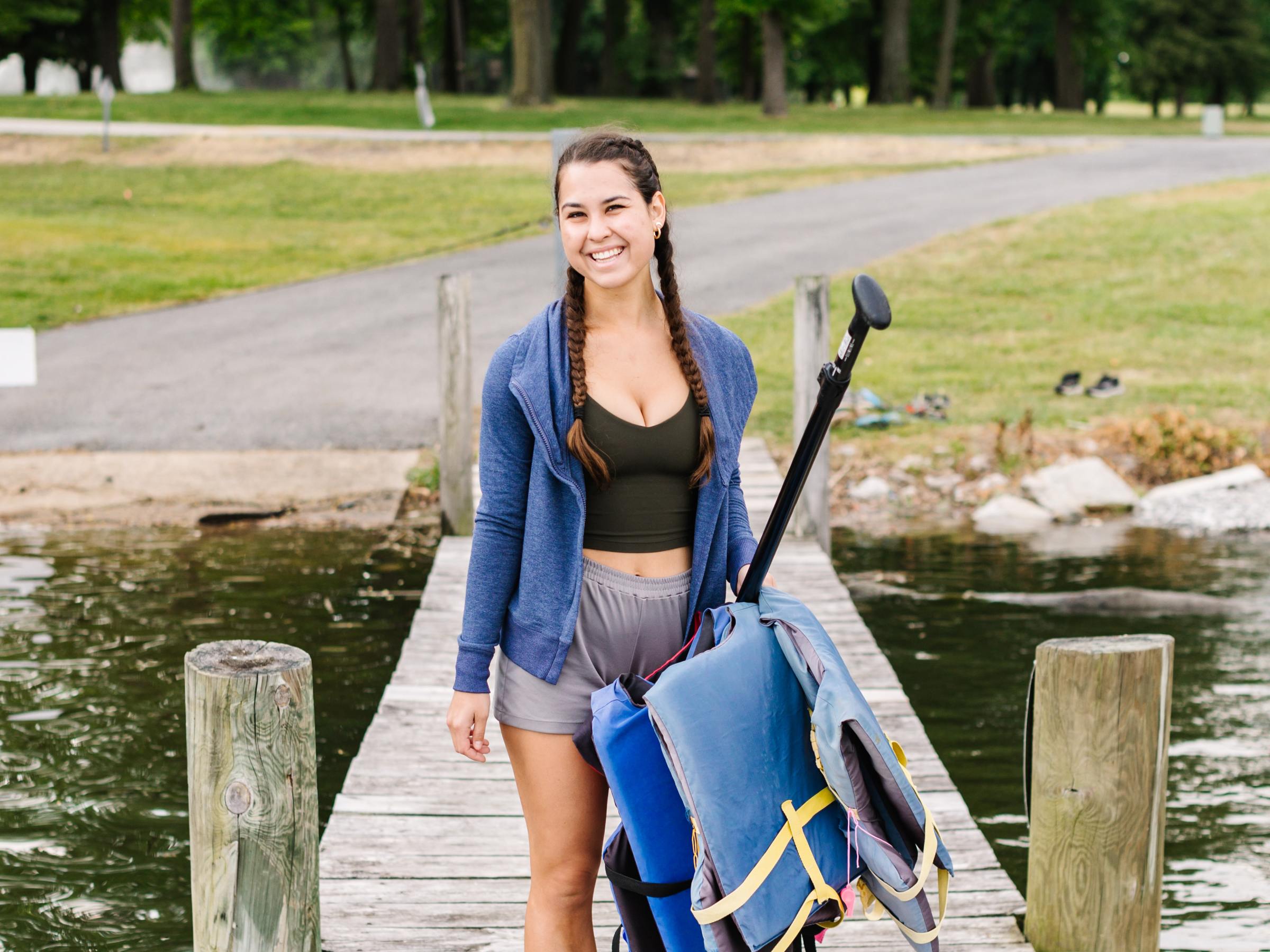 Woman with braids carrying paddle gear on a dock by a wooded lakeside.