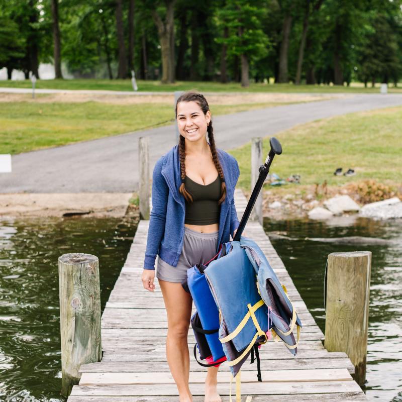 Woman with braids carrying paddle gear on a dock by a wooded lakeside.
