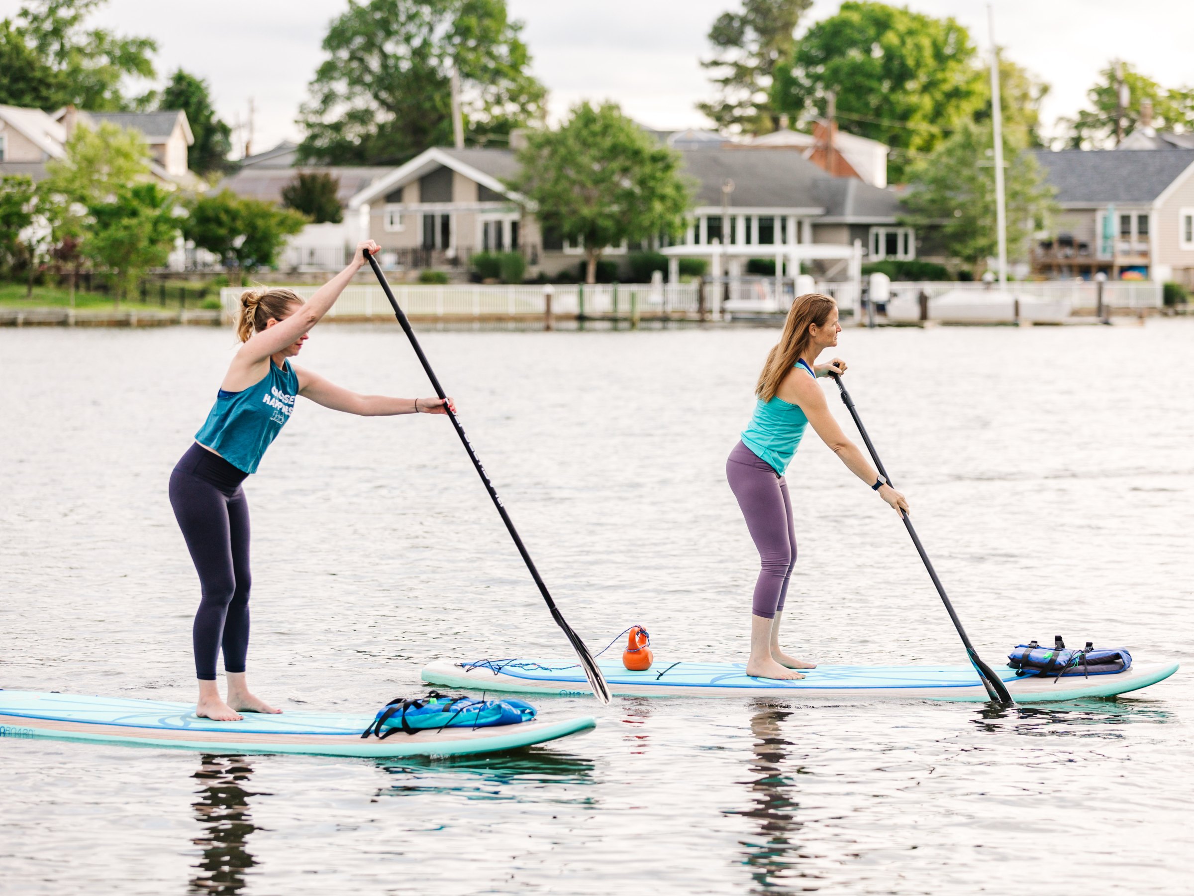 Two women paddleboarding on a calm lake near houses.