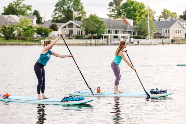 Two women paddleboarding on a calm lake near houses.