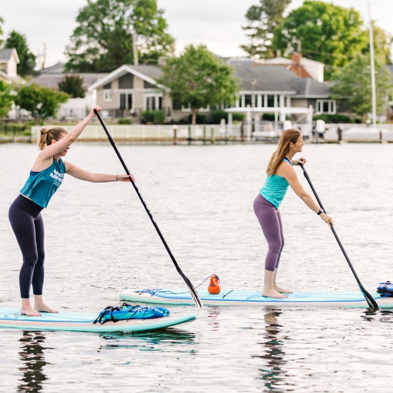 Two women paddleboarding on a calm lake near houses.