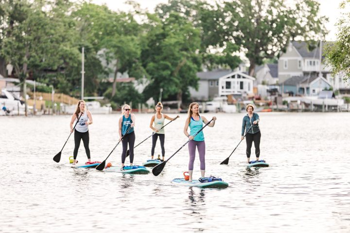 Five women paddleboarding on a calm lake near houses and trees on the shore.