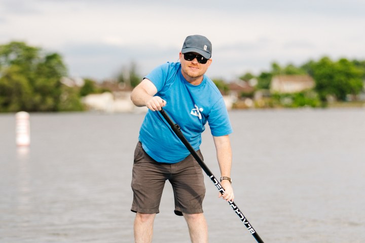 Man paddleboarding on calm water, wearing blue shirt and cap.
